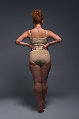 back view of curvaceous redhead woman in beige lingerie with hands on waist on dark grey backdrop clipart