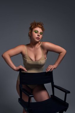 redhead plus size model in taupe underwear posing with chair on dark grey backdrop, body confidence clipart