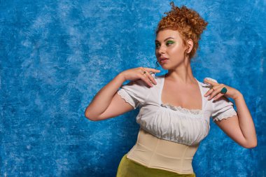 fashionable and redhead plus size woman in white blouse looking away on blue textured backdrop clipart