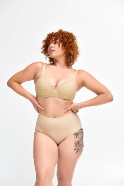 curvy and redhead tattooed model in beige lingerie with hands on hips looking away on white clipart