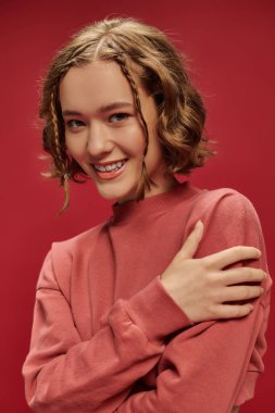 Kısa saçlı, örgülü, kırmızı arka planda kendine sarılan neşeli genç bir kadının portresi.