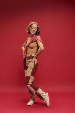 Kırmızı arkaplanda uzun kollu kazakla poz veren yama pantolonlu genç bir kadın.