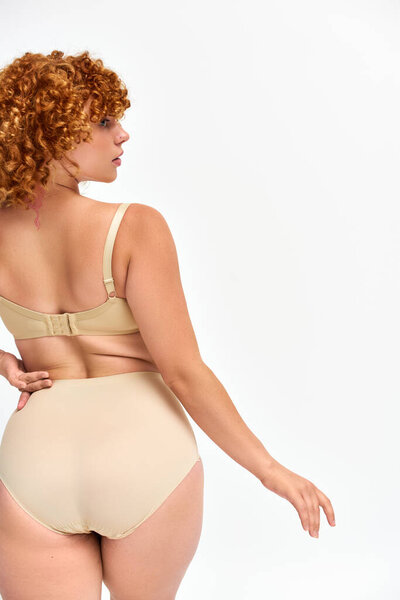 back view of redhead plus size woman in lingerie standing with hand on hip and looking away on white