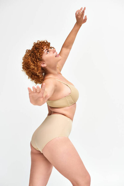 happy plus size redhead woman in beige lingerie standing and looking up in expressive pose on white