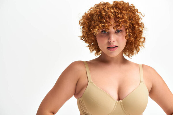 portrait of charming woman with red curly hair and curvy body and bust looking at camera on white
