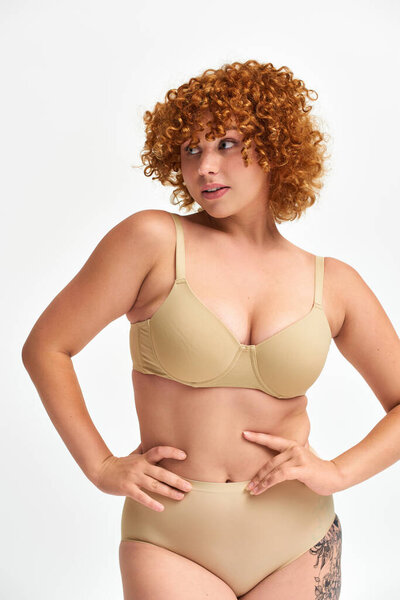очаровательная рыжая кривая модель в бежевом белье позирует с руками на бедрах и смотрит в сторону на белый