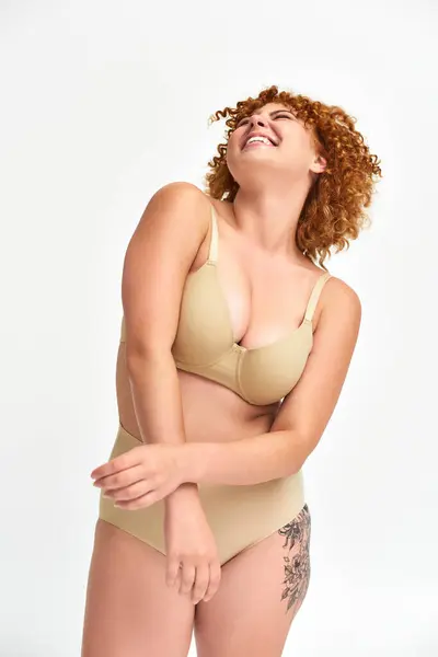 excited tattooed redhead woman with curvy body laughing with closed eyes on white, beige lingerie
