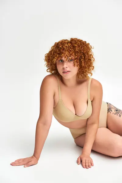 stock image seductive tattooed woman with red curly hair and plus size body looking at camera on white
