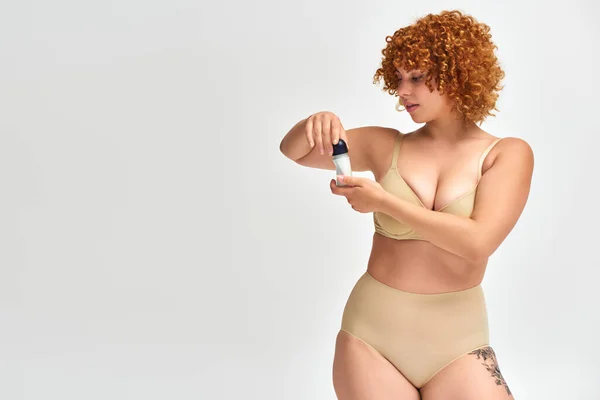 stock image curvy woman in lingerie with red wavy hair opening roll-on antiperspirant on white, self-care ritual