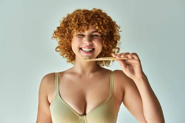 stock image cheerful redhead and curvy woman with toothbrush smiling with closed eyes on beige backdrop