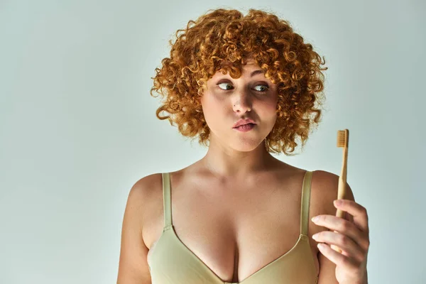 discouraged and redhead plus size woman in beige bra looking at toothbrush on grey background