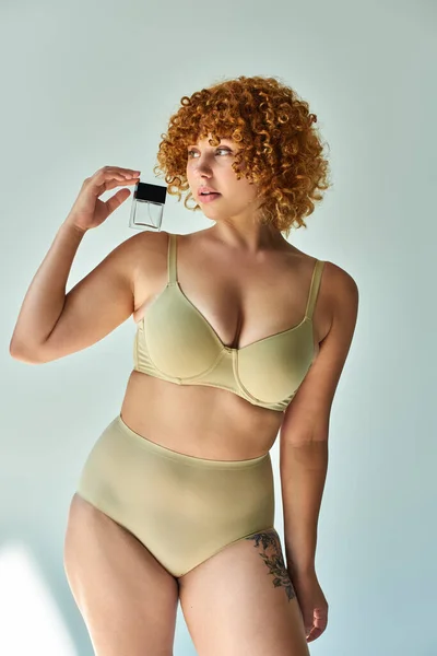 young curvy model in beige lingerie with red wavy hair holding perfume and looking away on grey