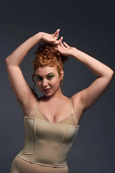 smiling curvy woman with red hair and bold makeup posing with hands above head on dark grey