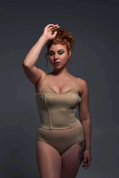 young plus size model with curvy body in beige underwear standing with hand near head on dark grey