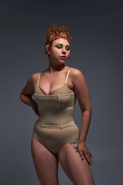 young redhead plus size woman with bold makeup posing in taupe underwear on dark grey backdrop