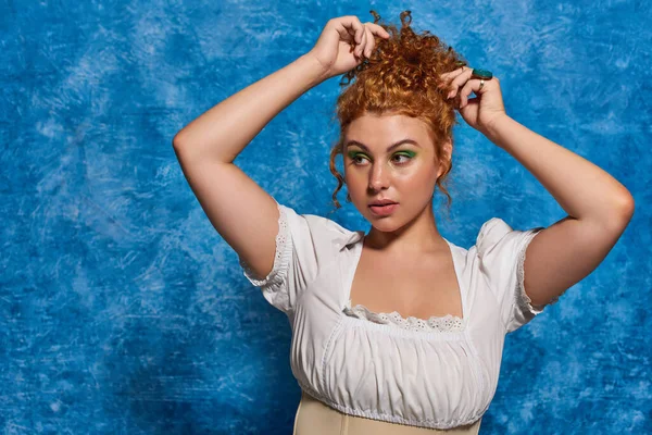 stock image stylish curvy woman in white blouse adjusting red hair on blue textured backdrop, plus size fashion