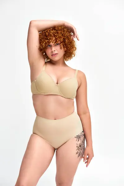 sensual plus size tattooed model in beige lingerie with hands above head looking at camera on white