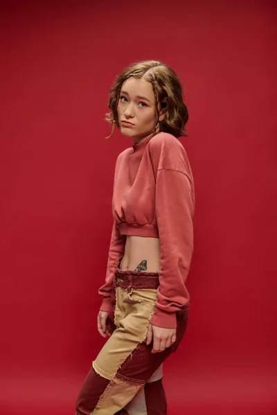 innocent look, young woman in patchwork pants and cropped long sleeve posing on red backdrop
