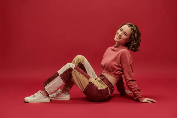stock image self-expression, positive woman in patchwork pants and cropped long sleeve sitting on red backdrop