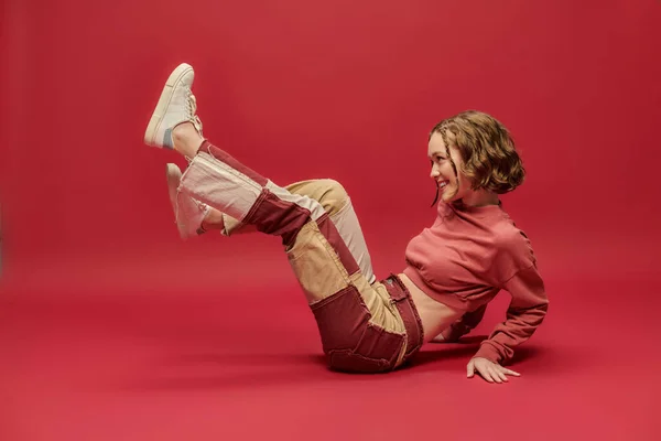 stock image self-expression, joyful woman in patchwork pants and cropped long sleeve sitting on red backdrop
