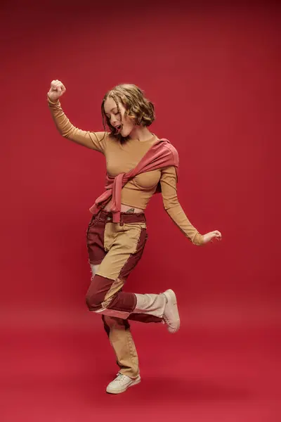 穿着拼凑裤的快乐的年轻女子在红色背景的裁剪长袖上摆出跳跃姿势 — 图库照片