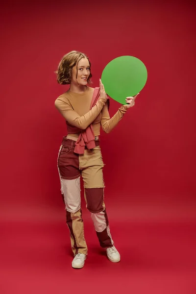 young woman in patchwork pants and jumper over cropped long sleeve pointing at speech bubble on red