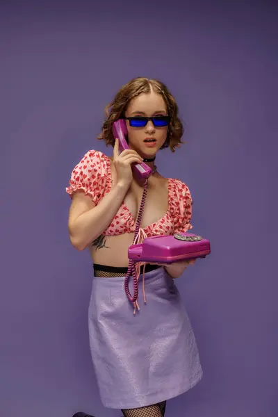 stock image pretty girl in trendy sunglasses and cropped top with hearts talking on retro telephone on purple