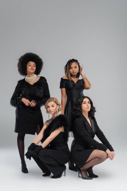 Sarışın ve esmer kız arkadaşlar, zarif Afro-Amerikan kadınlarının yanında gri popolarda poz veriyorlar.