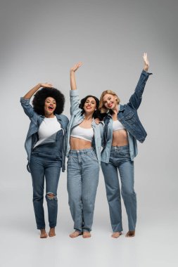 full length of joyful barefoot multiethnic girlfriends in blue denim wear waving hands on grey clipart