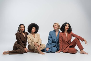 Gri arka planda oturan çok renkli takım elbiseli ve çıplak ayaklı çok ırklı kız arkadaşlar.