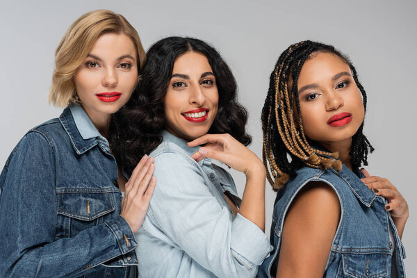 portrait of multiracial girlfriends in blue denim wear looking at camera on grey, diverse beauty