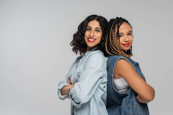 stylish multiracial women in denim outfits standing back to back with folded arms on grey, fashion