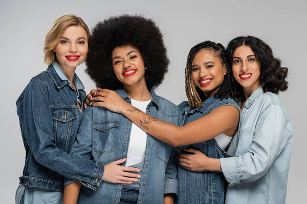 jolly and trendy multicultural girlfriends in blue denim wear smiling at camera on grey, friendship