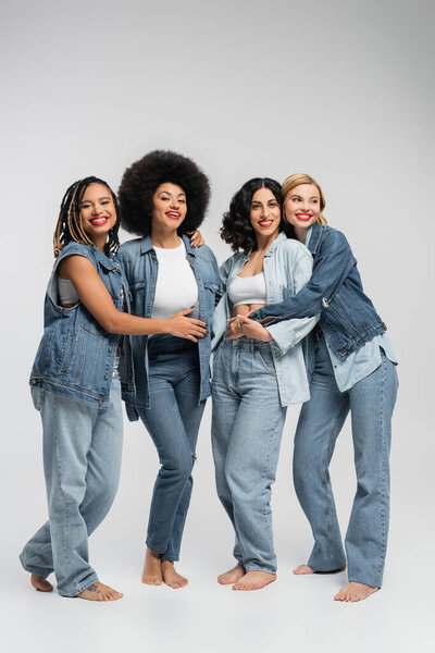smiley and barefoot multiethnic women in blue denim attire embracing on grey, diverse friendship