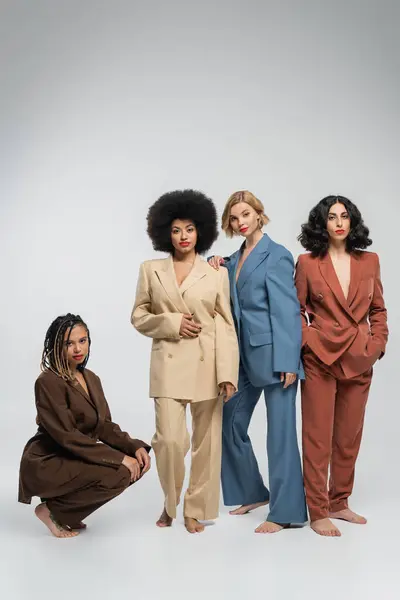 full length of multiracial barefoot girlfriends in colorful suits on grey backdrop, diverse beauty