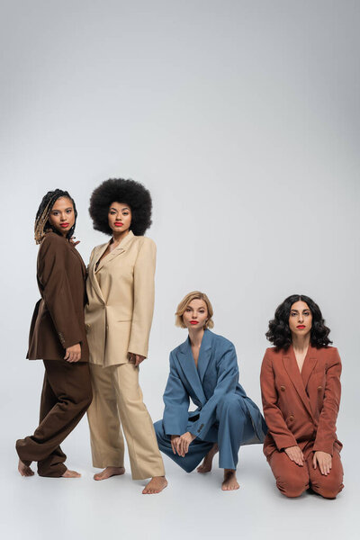 diverse beauty, barefoot multiracial girlfriends in colorful suits looking at camera on grey