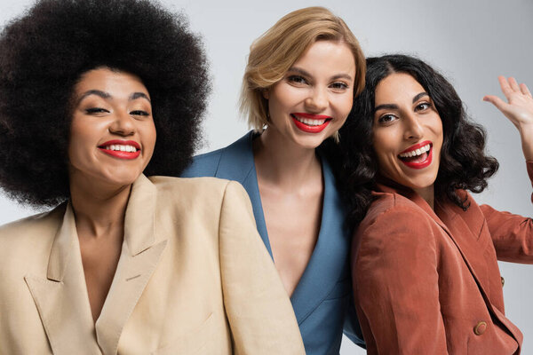 cheerful multiracial woman waving hands near smiley female friends in colorful suits on grey