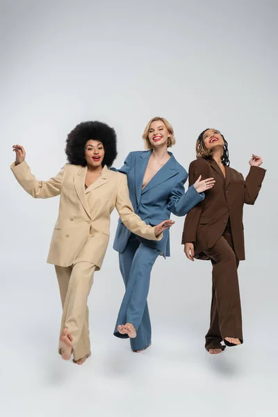 joyful barefoot multiracial fashion models in colorful suits posing barefoot on grey, full length