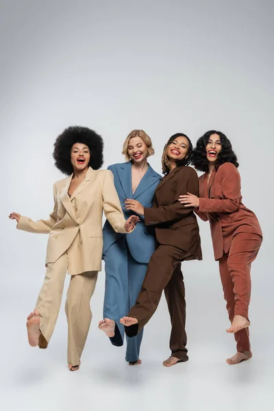 carefree barefoot multiracial girlfriends in multicolored suits posing barefoot on grey, full length