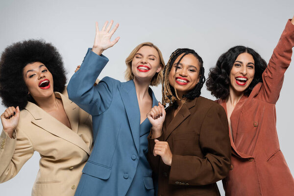 overjoyed multiracial girlfriends in colorful suits waving hands on grey, fashion and happiness