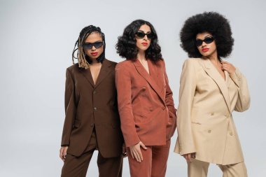 Renkli takım elbiseli, koyu güneş gözlüklü göz kamaştırıcı çok ırklı kız arkadaşlar gri, çeşitlilik üzerine poz veriyorlar.