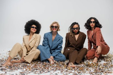 Güneş gözlüklü ve takım elbiseli modaya uygun çok ırklı kızlar gri renkli konfetiler üzerinde oturuyor, parti