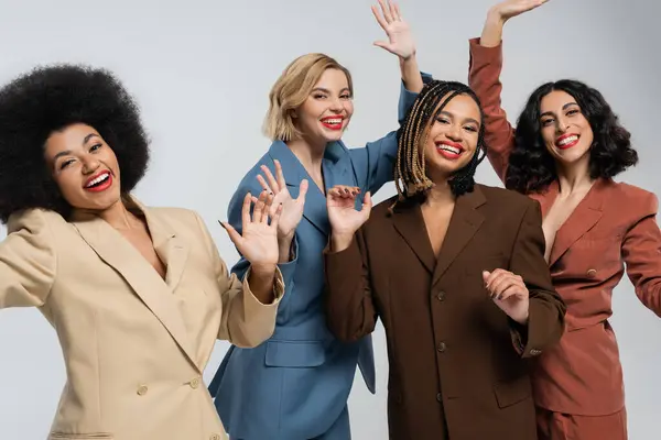 stock image inspired multiethnic girlfriends in multicolored suits waving hands on grey, beauty and diversity