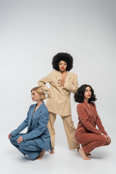 african american woman looking at camera near multiethnic girlfriends sitting on haunches on grey