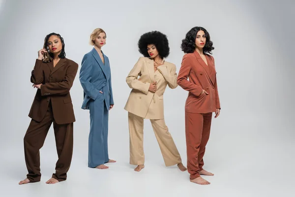 full length of multiracial barefoot female friends in trendy multicolored suits standing on grey
