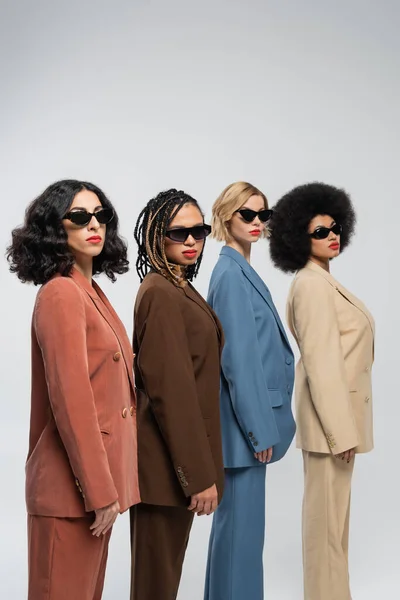 beauty in diversity, multiracial women in sunglasses and colorful suits looking at camera on grey