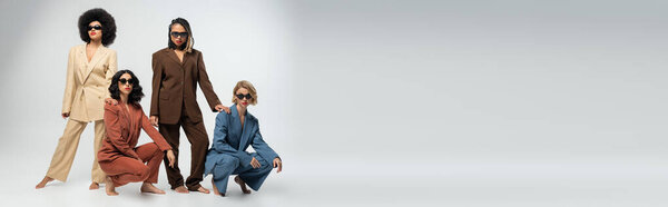 trendy group of barefoot multiethnic girlfriends in colorful suits and sunglasses on grey, banner