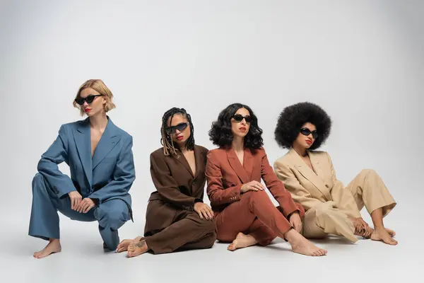 diverse fashion, barefoot multiethnic girlfriends in sunglasses and colorful suits sitting on grey