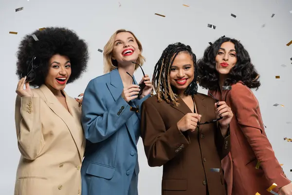 Renkli konfeti altında güneş gözlüğü takan şık takım elbiseli çok ırklı kızlar, parti