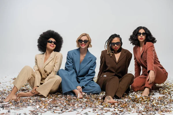 Güneş gözlüklü ve takım elbiseli modaya uygun çok ırklı kızlar gri renkli konfetiler üzerinde oturuyor, parti